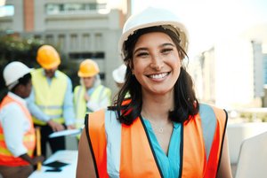 Eine junge weibliche Bauarbeiterin mit Warnweste und Schutzhelm lächelt in die Kamera. Im Hintergrund sind 3 weitere Arbeiter*innen.